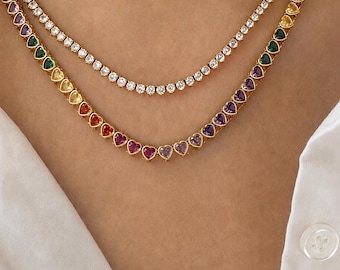 May include: Two layered necklaces. The top necklace is a gold-tone chain featuring a continuous row of clear, round-cut stones. The bottom necklace is a gold-tone chain featuring a continuous row of heart-shaped stones in various rainbow colors.