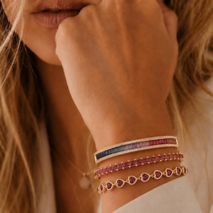 May include: A stack of three gold bracelets. The top bracelet features a gradient of baguette-cut stones transitioning from dark blue to pink. The middle bracelet has square-cut pink stones. The bottom bracelet consists of linked gold hearts.