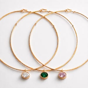 May include: Three gold-toned hoop necklaces with gemstone pendants. One pendant features a clear stone, another a green stone, and the third a pink stone. The necklaces are displayed on a white background.