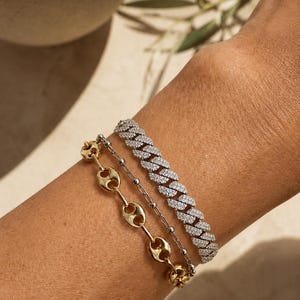 May include: A set of three bracelets worn together. One is a gold-toned chain with oval links, the second is a thin silver-toned chain with small spherical beads, and the third is a silver-toned cuban link chain encrusted with clear stones.