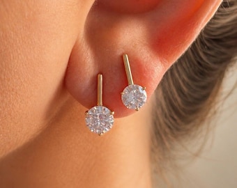 May include: A pair of gold earrings featuring a vertical bar design attached to a round, clear, faceted gemstone. The earrings are positioned on an earlobe, showcasing the metallic gold finish and the reflective, brilliant cut of the stones.