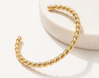May include: A gold-colored, open cuff bracelet with a twisted rope design. The bracelet is made of a shiny, metallic material and is displayed on a white surface. The bracelet is a piece of jewelry.