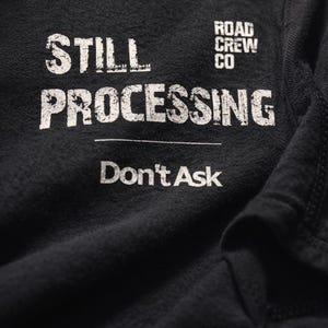 May include: Close-up of a black t-shirt with white text. The text reads "STILL PROCESSING" above "Don't Ask". The words "ROAD CREW CO" are printed above the word "PROCESSING".