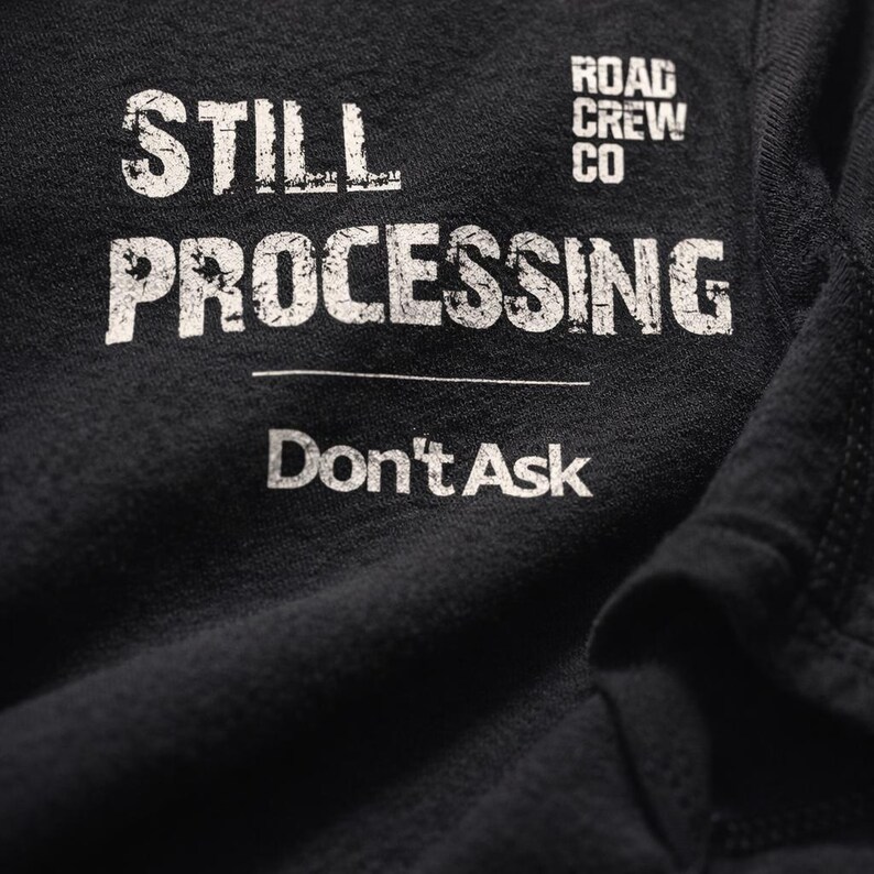 May include: Close-up of a black t-shirt with white text. The text reads "STILL PROCESSING" above "Don't Ask". The words "ROAD CREW CO" are printed above the word "PROCESSING".