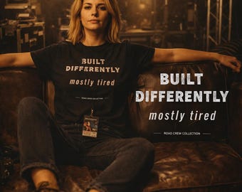 May include: A woman wearing a black t-shirt with the text "BUILT DIFFERENTLY mostly tired" sitting on a brown leather couch. The shirt is part of the Road Crew Collection. She is wearing dark jeans and brown boots.