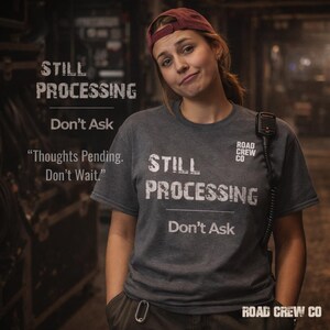 May include: A woman wearing a gray t-shirt with the words "STILL PROCESSING Don't Ask" printed on the front. The shirt also features the text "Thoughts Pending. Don't Wait." and the Road Crew Co. logo. She is wearing a maroon baseball cap and holding a microphone.
