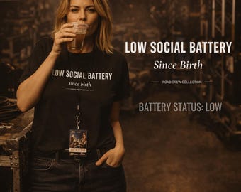 May include: A woman wearing a black t-shirt with the text "LOW SOCIAL BATTERY Since Birth" drinks from a clear glass. The image also includes the text "BATTERY STATUS: LOW" and "ROAD CREW COLLECTION".