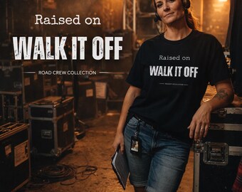 May include: A woman wearing a black t-shirt with the text "Raised on WALK IT OFF" in white, part of the Road Crew Collection. She is in a backstage setting, wearing headphones and denim shorts, holding a clipboard.