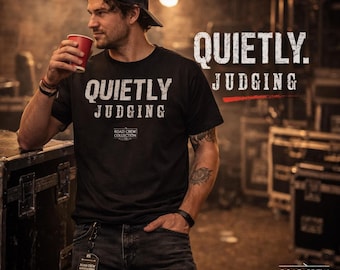 May include: Black t-shirt with the text "QUIETLY. JUDGING" and "ROAD CREW COLLECTION" in white. The image shows a person wearing the shirt, holding a red cup, and wearing a black cap and jeans. The background is a dimly lit stage.
