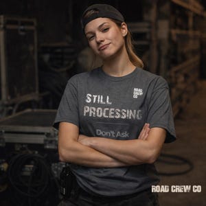 May include: A dark gray t-shirt with the words "STILL PROCESSING" and "Don't Ask" printed in white. The shirt also features the text "ROAD CREW CO" in white. The person in the image is wearing a black cap.
