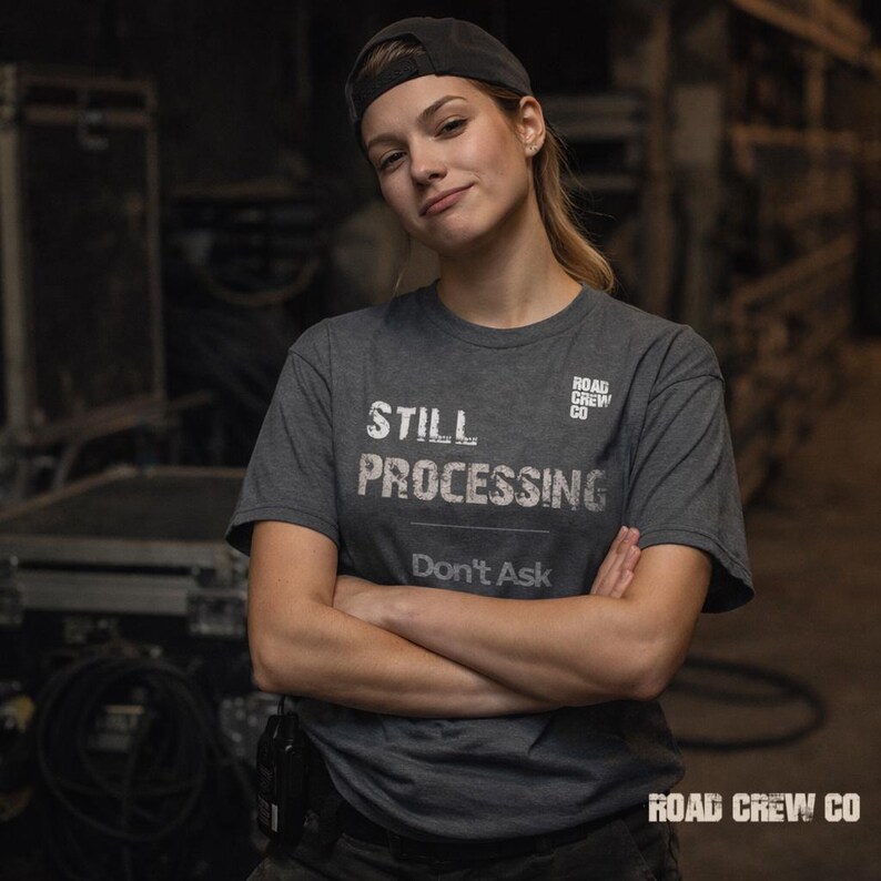 May include: A dark gray t-shirt with the words "STILL PROCESSING" and "Don't Ask" printed in white. The shirt also features the text "ROAD CREW CO" in white. The person in the image is wearing a black cap.