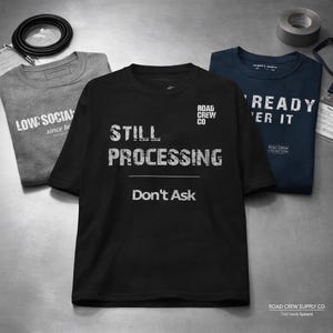 May include: Three t-shirts with text. A black t-shirt reads "STILL PROCESSING Don't Ask". A gray t-shirt reads "LOW: SOCIAL since bi". A navy blue t-shirt reads "READY 'ER IT". The shirts are from Road Crew Supply Co.