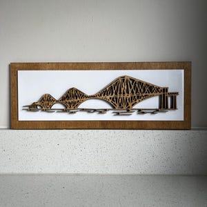 May include: A framed wooden artwork showing a laser-cut representation of a bridge. The bridge is crafted from light brown wood, set against a white backdrop. The frame is a darker brown. The rectangular artwork is likely a decorative item for a home.