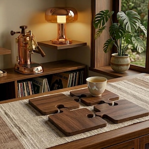 Puede incluir: Cuatro posavasos de madera con forma de rompecabezas sobre una mesa con un camino de mesa tejido de color claro. Una taza de té está sobre un posavasos. Una máquina de espresso de cobre y una lámpara retro están en el fondo.