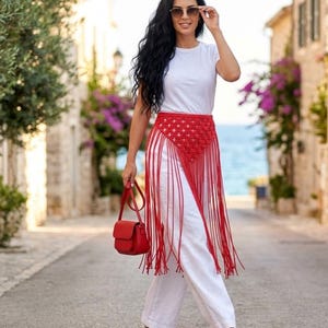 May include: A white short-sleeved t-shirt and white wide-leg trousers paired with a red macrame waist belt featuring a diamond-patterned weave and long hanging fringe. A small red structured handbag and white open-toe sandals with red accents.
