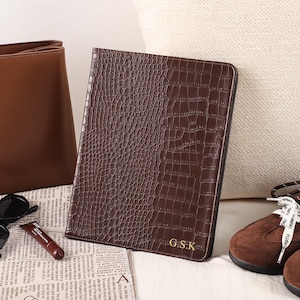 Peut inclure: Une couverture de carnet rectangulaire marron foncé avec un motif texturé en peau de crocodile. Le coin inférieur droit présente les initiales G.S.K. gaufrées en or. La couverture est dotée de bords cousus et d'un profil plat et fin.