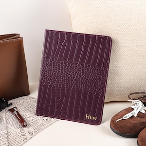 May include: A rectangular dark purple notebook with a textured crocodile skin pattern and the name Huw embossed in gold lettering on the bottom right corner. The notebook features stitched edges and a slim, upright design.