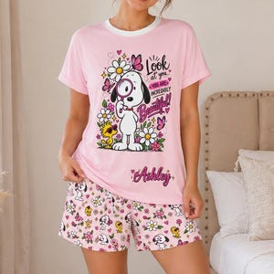 May include: A light pink pajama set consisting of a short-sleeved t-shirt and shorts. The shirt features a graphic of Snoopy holding a magnifying glass with the text Look at you, YOU ARE INCREDIBLY Beautiful and the name Ashley in cursive script.