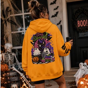 Puede incluir: Una sudadera con capucha de color naranja brillante que presenta un gran gráfico en la espalda con el texto Stay Spooky. El diseño incluye dos fantasmas, una calabaza, una pila de libros, una taza de calavera y una casa encantada.
