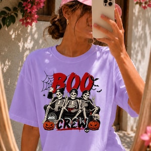Puede incluir: Una camiseta de manga corta de color morado claro con un estampado gráfico de tres esqueletos sentados en un sofá. El texto dice BOO CREW en rojo y negro. El diseño incluye una telaraña, murciélagos y dos calabazas naranjas en la parte inferior.