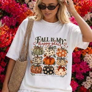 Puede incluir: Una camiseta blanca de manga corta con un gráfico de nueve calabazas estilizadas con varios patrones, incluyendo cuadros, flores y estrellas. El texto dice Fall is my happy place, crisp air y pumpkin season en verde oscuro y naranja.