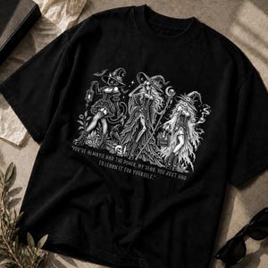 Puede incluir: Una camiseta negra con un estampado gráfico blanco de tres figuras tipo bruja con sombreros, incluyendo un personaje felino, un esqueleto y una mujer, con el texto: "YOU'VE ALWAYS HAD THE POWER, MY DEAR. YOU JUST HAD TO LEARN IT FOR YOURSELF."