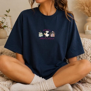 May include: Navy blue t-shirt with a whimsical "Book Club" design. The graphic features a goose wearing a floral crown, holding a book, and standing between stacks of books and flowers. The text "Addicted to good stories" is below the graphic.