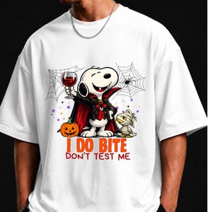 Peut inclure: Un t-shirt blanc avec un motif représentant un chien de dessin animé déguisé en vampire, tenant un verre de liquide rouge, à côté d'une citrouille et d'une momie. Le texte indique I DO BITE DON'T TEST ME, entouré de toiles d'araignée.