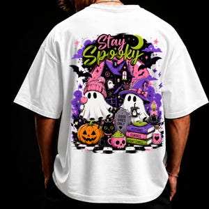 Puede incluir: Una camiseta blanca con un gráfico colorido en la espalda. El diseño incluye dos fantasmas, uno con un gorro rosa y otro con un sombrero de bruja morado, un gato negro, una calabaza, libros etiquetados como Potions, Curses, Spooky Stories y el texto Stay Spooky y Good Vibes Only.