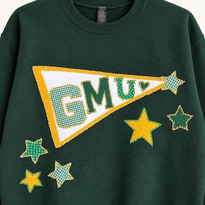May include: A dark green crewneck sweatshirt featuring a large white pennant applique with yellow trim and the letters GMU in green and white gingham. The design includes several yellow and green gingham star-shaped fabric appliques.
