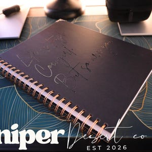 May include: A black spiral-bound notebook with gold accents and the inscription "Life is tough, but so are you." The notebook is on a patterned surface with the Juniper Desert co. logo.