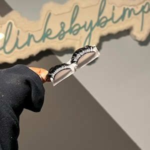 Puede incluir: Un par de pestañas postizas en blanco y negro sobre un respaldo blanco. Las pestañas se sostienen frente a un letrero que dice "linksbybimp" en escritura cursiva.