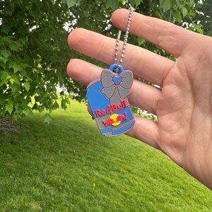 May include: A blue and gray Red Bull keychain with a silver chain. The keychain features a bow and the Red Bull logo in red and yellow. The background is a green lawn and foliage.