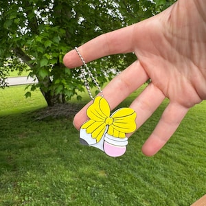 Puede incluir: Un collar con colgante colorido con un dise&ntilde;o de l&aacute;piz con un lazo amarillo, una goma de borrar rosa y una punta gris. El colgante cuelga de una cadena plateada. El fondo es un c&eacute;sped verde y un &aacute;rbol.