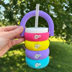 May include: A colorful, stackable baby bottle with a white lid and a clear straw. The bottle is made of silicone and features a purple handle. The bottle is composed of pink, yellow, purple, and blue sections.