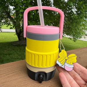 Puede incluir: Una jarra de agua aislada multicolor con asa y pajita rosas. La jarra es amarilla, gris, rosa, beige y negra. Un dije en forma de lazo amarillo cuelga del asa. La jarra tiene un grifo para dispensar l&iacute;quidos.