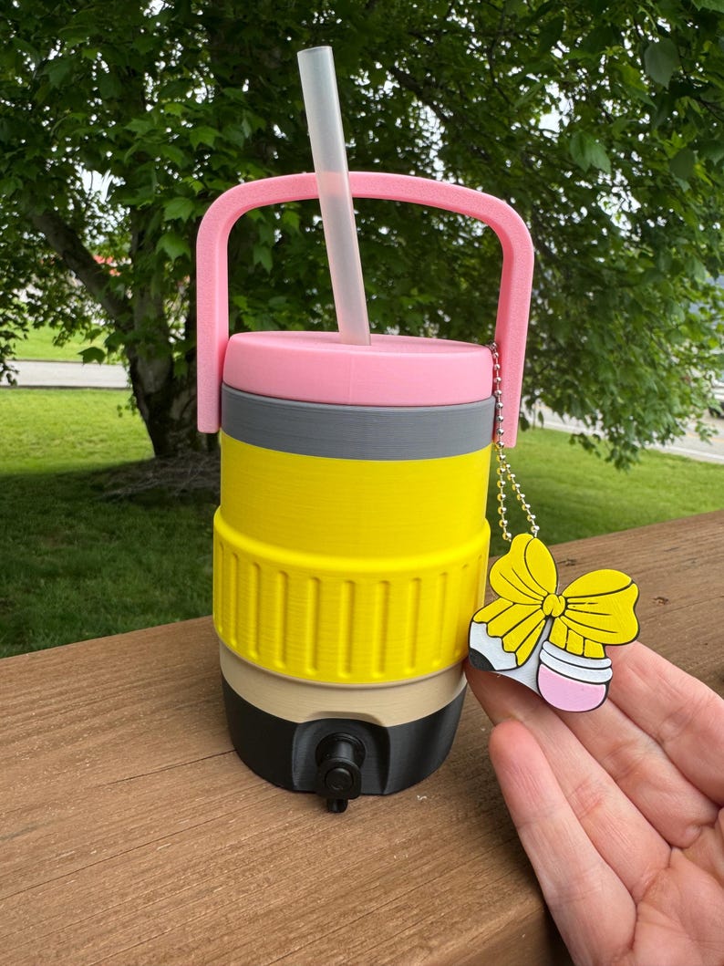 Puede incluir: Una jarra de agua aislada multicolor con asa y pajita rosas. La jarra es amarilla, gris, rosa, beige y negra. Un dije en forma de lazo amarillo cuelga del asa. La jarra tiene un grifo para dispensar l&iacute;quidos.