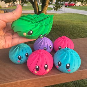 May include: A collection of five spherical, articulated toys with ribbed textures and cartoon faces. One green toy is held, while four others in blue, purple, and pink are arranged below. Each features large black eyes and small blush marks.