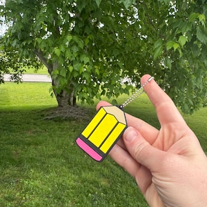 May include: A yellow pencil-shaped keychain with black and pink accents. The keychain is held up against a backdrop of green grass and a tree with lush green leaves. The pencil has a silver chain.
