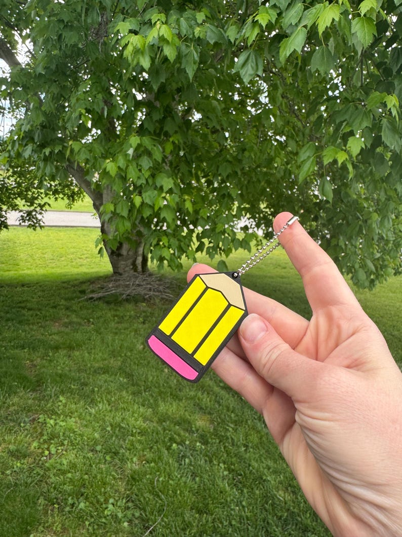 May include: A yellow pencil-shaped keychain with black and pink accents. The keychain is held up against a backdrop of green grass and a tree with lush green leaves. The pencil has a silver chain.