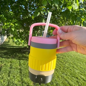 May include: A multi-colored insulated tumbler with a pink handle and straw. The tumbler is designed to resemble a pencil, with yellow, gray, tan, and black sections. The straw is white. The tumbler is held against a background of green grass and foliage.