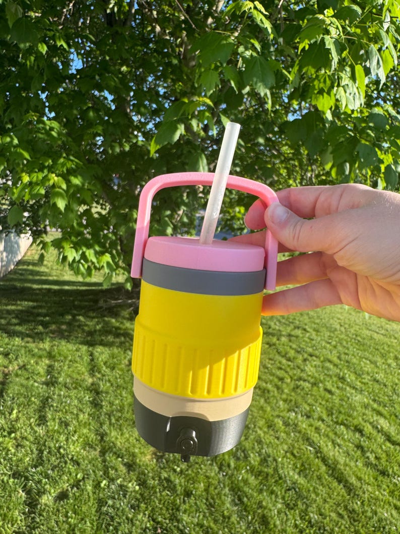 May include: A multi-colored insulated tumbler with a pink handle and straw. The tumbler is designed to resemble a pencil, with yellow, gray, tan, and black sections. The straw is white. The tumbler is held against a background of green grass and foliage.