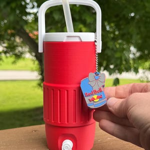May include: A red insulated beverage cooler with a white handle and lid, a white straw, and a silver chain with a Red Bull charm. The cooler has a spigot and is designed to keep drinks cold.