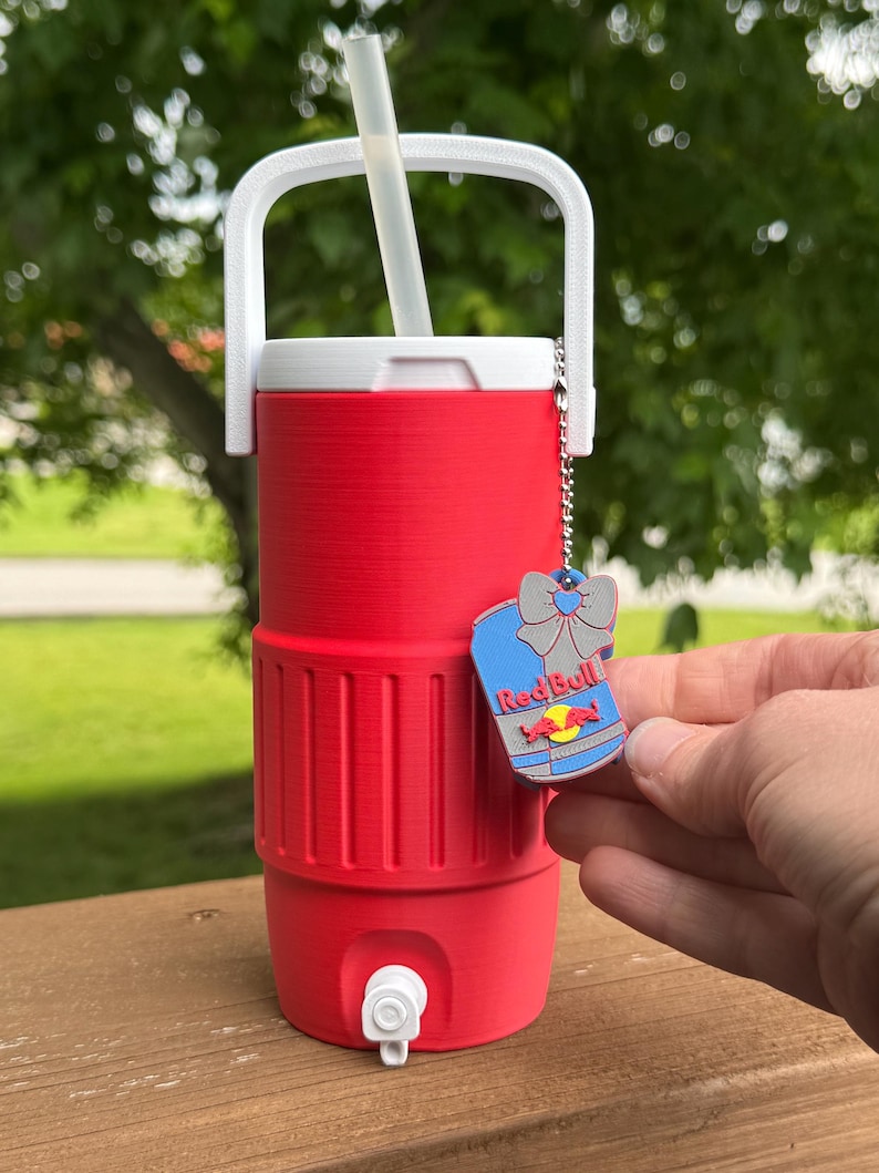 May include: A red insulated beverage cooler with a white handle and lid, a white straw, and a silver chain with a Red Bull charm. The cooler has a spigot and is designed to keep drinks cold.