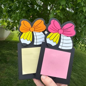 May include: Two black memo holders with pencil and bow designs. One has an orange bow and yellow sticky note, the other has a pink bow and pink sticky note. The background is a green, leafy tree.