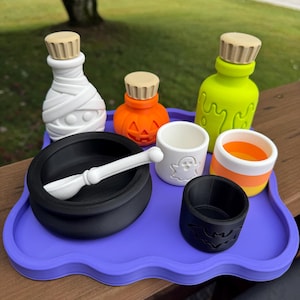 May include: A purple tray holding a white mummy-shaped bottle, an orange pumpkin-shaped bottle, a neon green bottle, a black cauldron with a white spoon, a white cup with a ghost, a black cup with a bat, and a candy corn-colored cup.