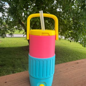 May include: A colorful beverage cooler with a yellow handle and lid, a pink midsection, and a turquoise base. A clear straw extends from the top. The cooler has a yellow spigot and is designed for outdoor use.