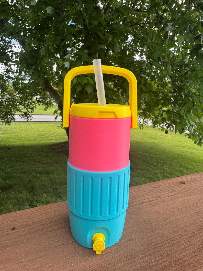 May include: A colorful beverage cooler with a yellow handle and lid, a pink midsection, and a turquoise base. A clear straw extends from the top. The cooler has a yellow spigot and is designed for outdoor use.