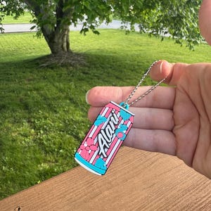 May include: A keychain featuring a colorful can design with the word "Alani" in white script. The can has a pink and white striped background with blue and pink floral accents. The keychain is attached to a silver chain.