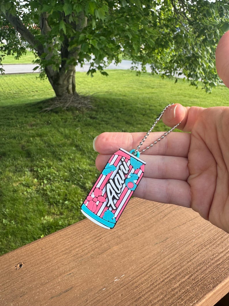 May include: A keychain featuring a colorful can design with the word "Alani" in white script. The can has a pink and white striped background with blue and pink floral accents. The keychain is attached to a silver chain.
